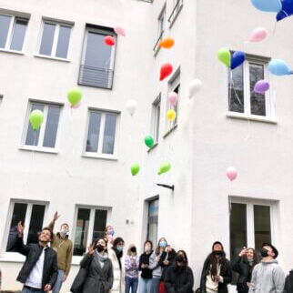 Menschen lassen bunte Luftballons vor einem weißen Gebäude in den Himmel steigen und feiern einen besonderen Moment.