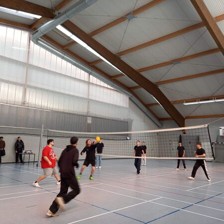 Mehrere Personen spielen Volleyball in einer großen Sporthalle mit hohen Decken und einem Netz in der Mitte.