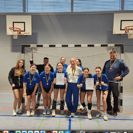 Eine Gruppe von jungen Athletinnen mit Medaillen posiert in einer Sporthalle vor einem Handballtor für ein Siegerfoto.