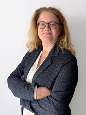 Eine Frau mit Brille und blonden Haaren trägt einen dunkel gestreiften Blazer und lächelt mit verschränkten Armen.