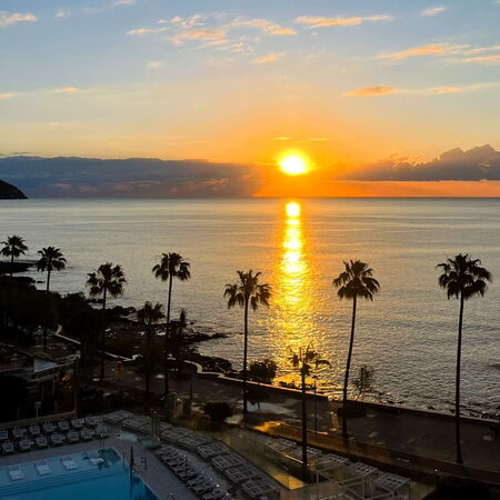 Die Sonne geht über dem Meer auf Mallorca unter, während Palmen und ein leerer Pool die friedliche Küstenszene rahmen.