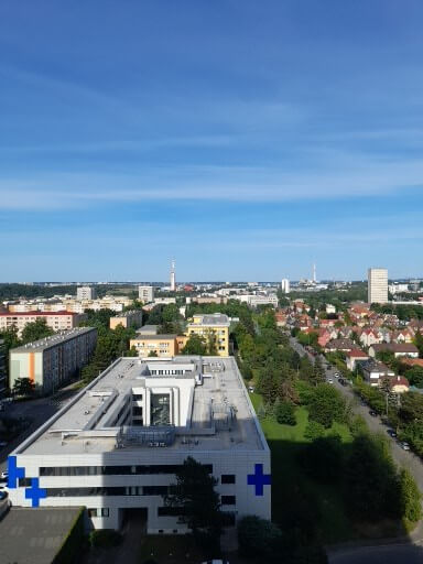 Blick über die Stadt mit modernen Gebäuden und blauen Kreuzsymbolen auf dem Krankenhausdach in der Ferne.