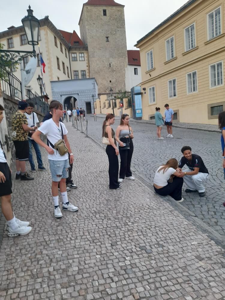 Eine Gruppe junger Menschen steht und sitzt auf einer gepflasterten Straße, umgeben von historischen Gebäuden.