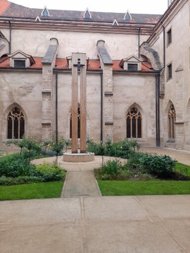 Ein ruhiger Innenhof mit einem Holzkreuz, umgeben von grünen Pflanzen und historischen Gebäuden.
