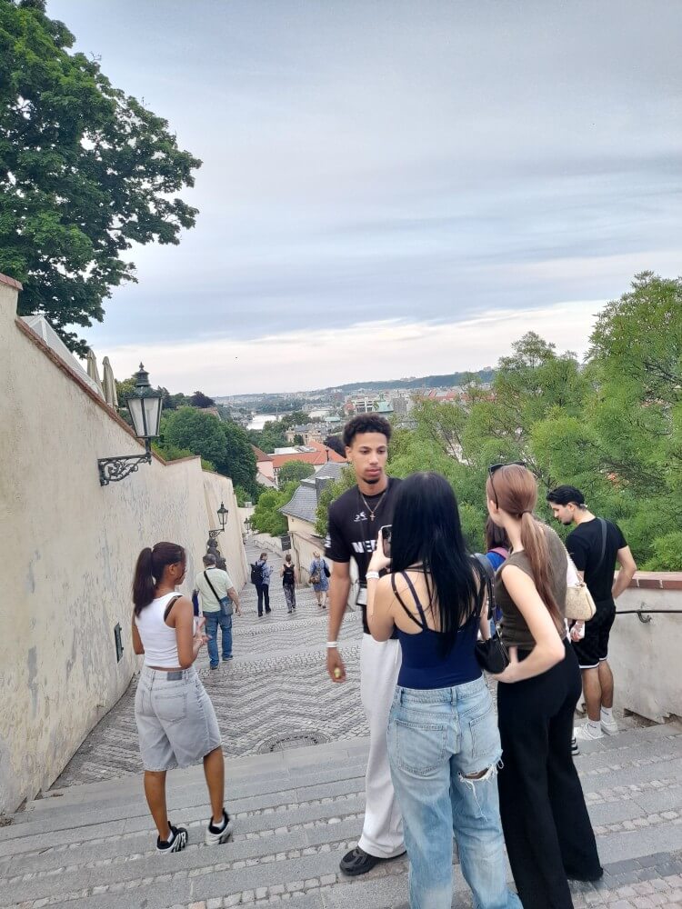 Freunde unterhalten sich auf einer Treppe mit Blick auf die Stadt, umgeben von Bäumen und einem bewölkten Himmel.