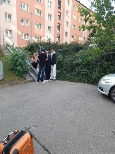 Eine Gruppe von Menschen sitzt und steht an einer Treppe vor einem Wohnblock, umgeben von Pflanzen und Fahrzeugen.