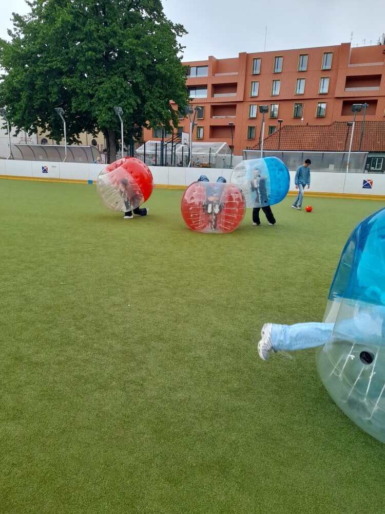 Kinder spielen auf einem Sportplatz in aufblasbaren Bällen, während ein Baum im Hintergrund steht.