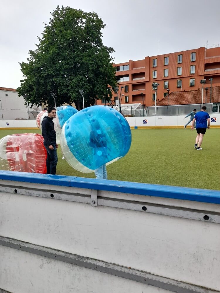 Zwei Personen in aufblasbaren Bällen stehen auf einem Sportplatz, während andere Spieler sich vorbereiten.