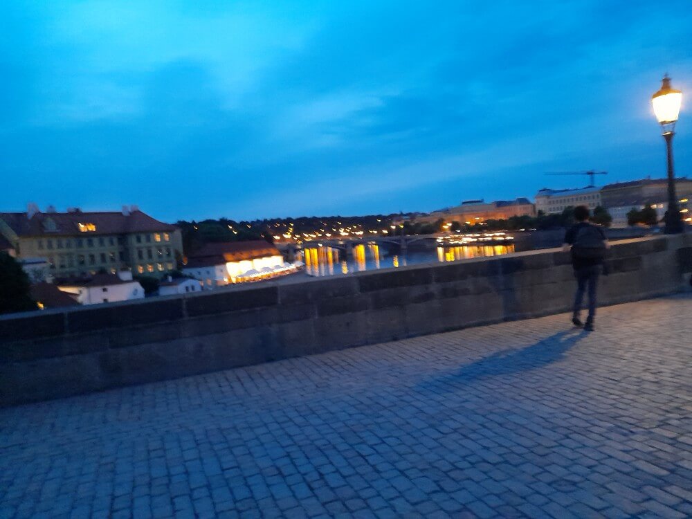 Ein Mensch steht an einer Steinmauer und blickt auf das beleuchtete Wasser und die Gebäude bei Dämmerung.