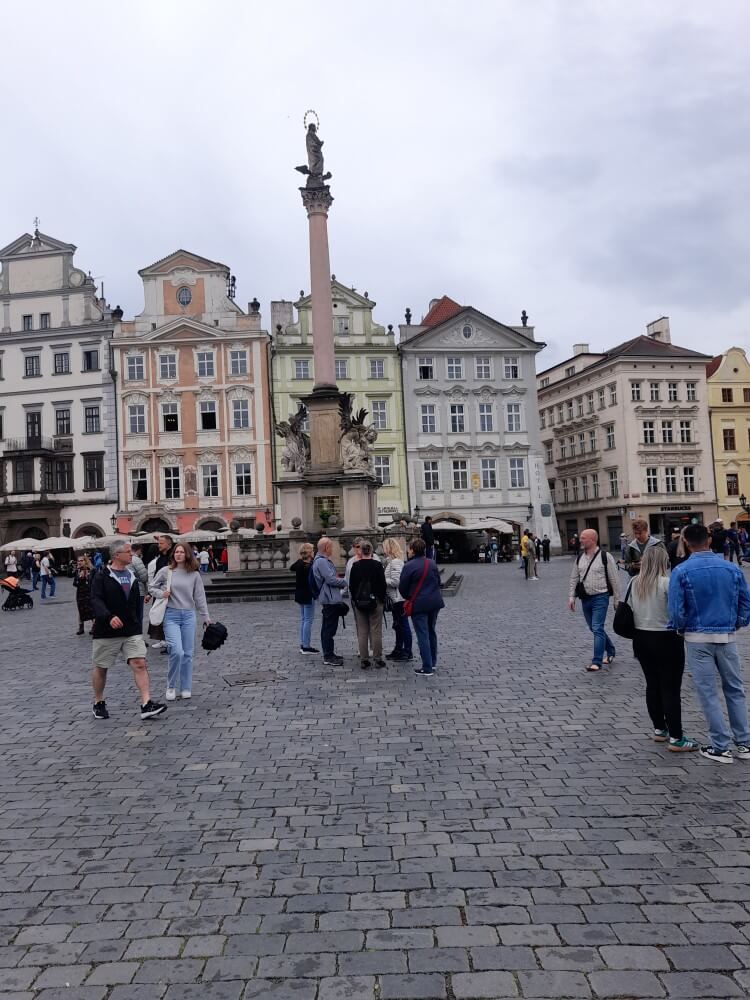 Menschen stehen auf einem Platz mit historischen Gebäuden und einer Statue in der Mitte, unter einem bewölkten Himmel.