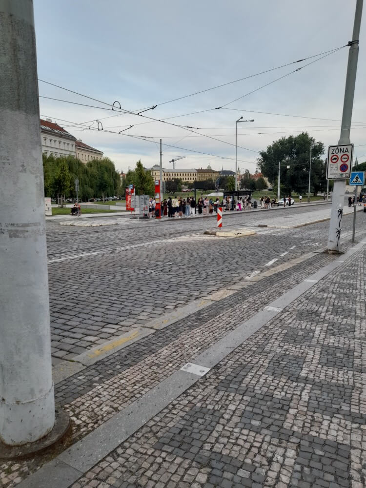 Menschen warten an einer Straßenbahnhaltestelle vor einem modernen Stadtbild mit Bäumen und Gebäuden.