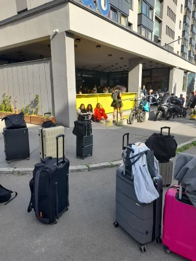 Reisende warten vor einem Hotel mit Gepäck, während andere am Eingang stehen und auf Einlass warten.