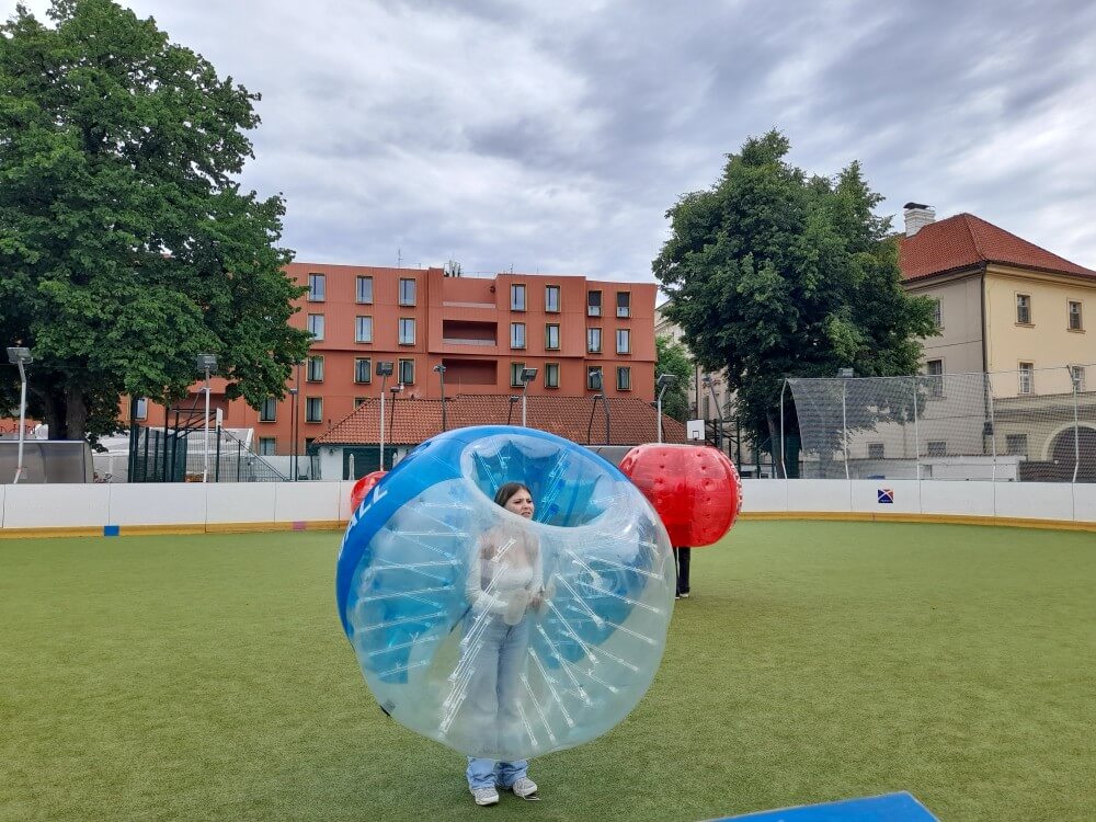 Eine Person steht in einem aufblasbaren Ball auf einem Sportfeld mit Bäumen und Gebäuden im Hintergrund.