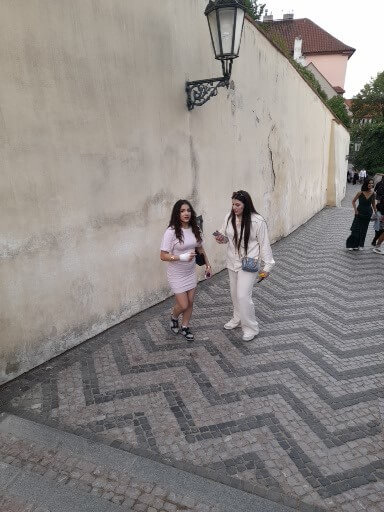 Zwei Frauen spazieren auf einem gepflasterten Weg entlang einer alten Mauer in einer malerischen Umgebung.