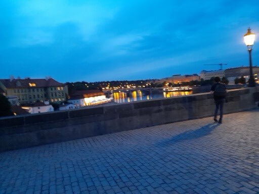 Eine Person steht auf einer Brücke, während die Stadt bei Abenddämmerung und reflektierendem Wasser erleuchtet ist.
