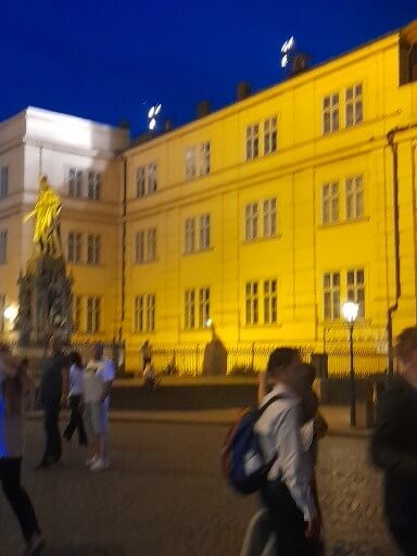 Beleuchtetes gelbes Gebäude mit Statue, abends auf einem Platz, umgeben von Menschen und Laternen.
