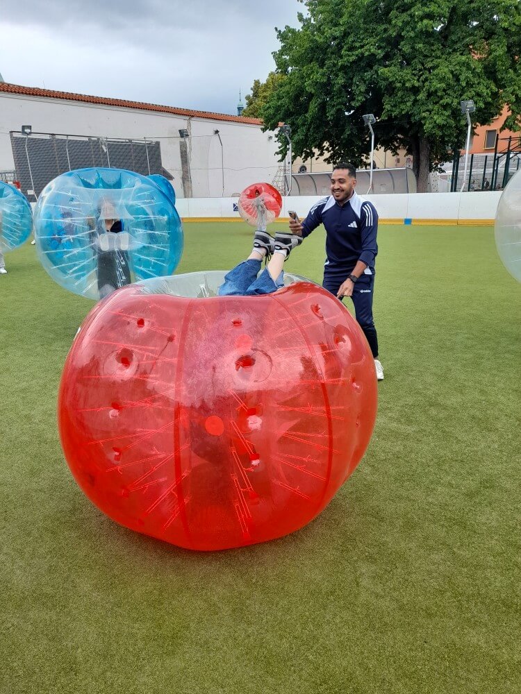 Menschen spielen Bubble Soccer in farbigen, aufblasbaren Bällen auf einem Rasenfeld, lachen und machen Fotos.