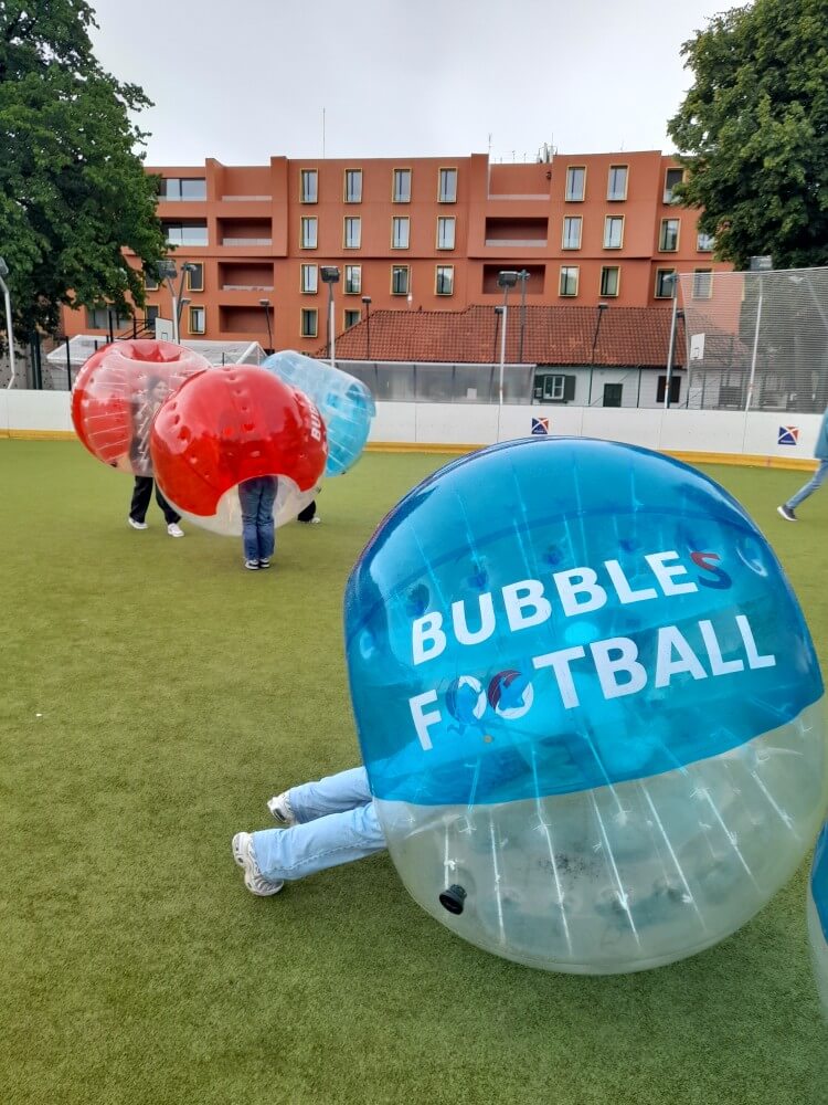 Kinder spielen Bubble Football in bunten, transparenten Bubbles auf einem Rasenfeld unter bewölktem Himmel.