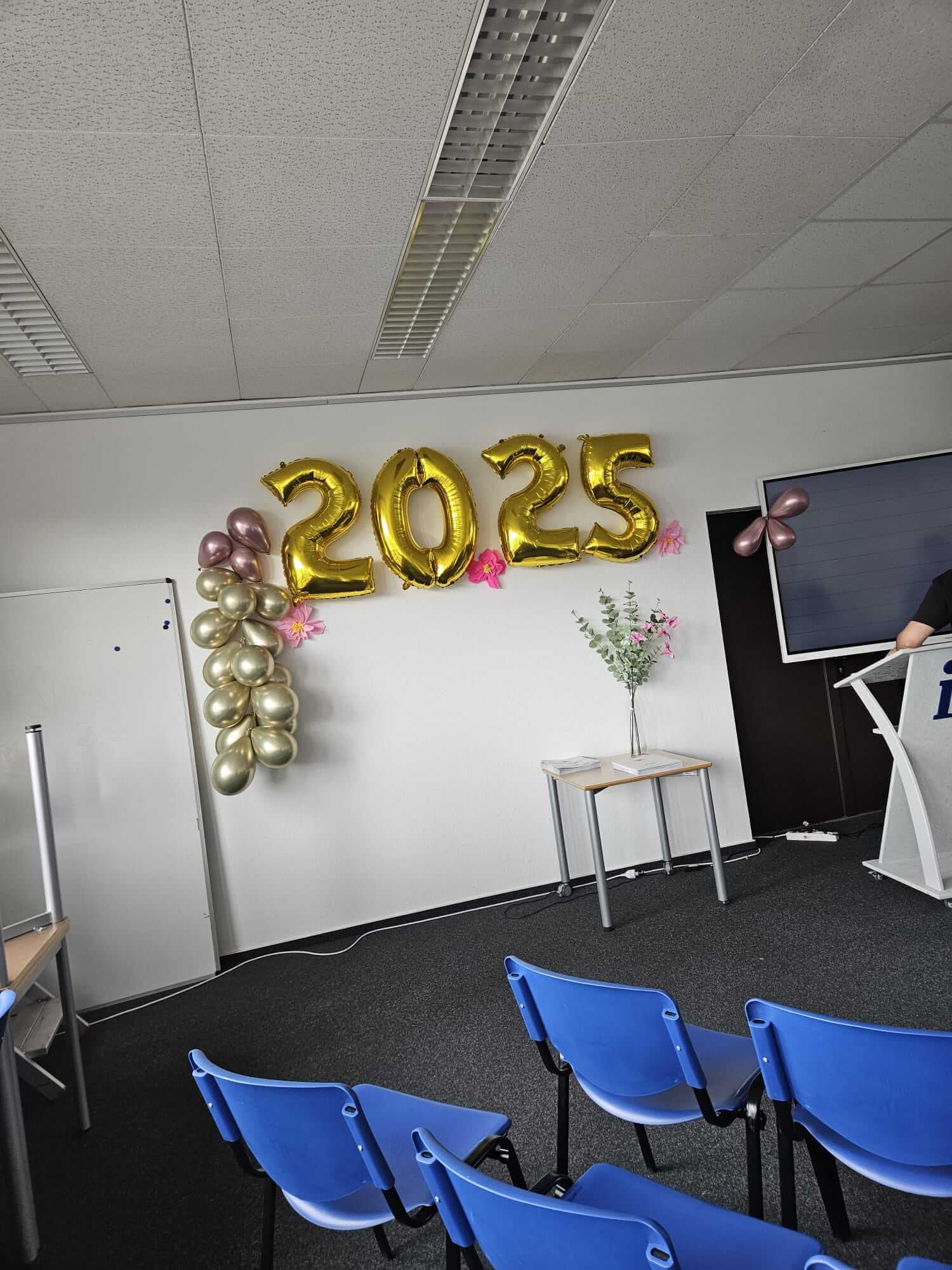 Ein Konferenzraum ist mit goldenen Luftballons in der Form von "2025" und blauen Stühlen dekoriert.
