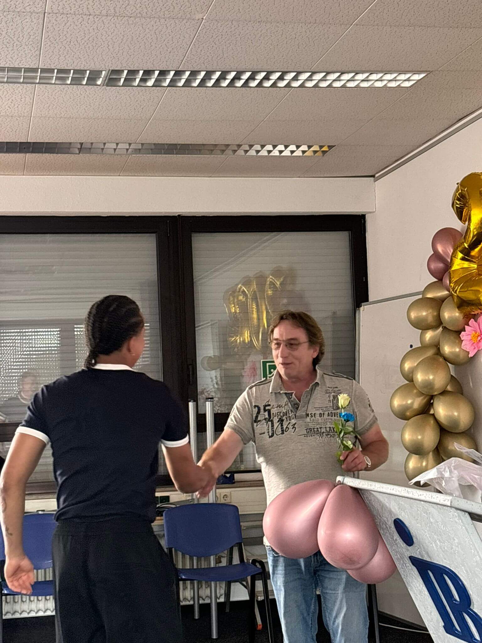Zwei Männer schütteln sich die Hand in einem Raum mit goldenen und rosafarbenen Luftballons bei einer Feier im Juli 2025.