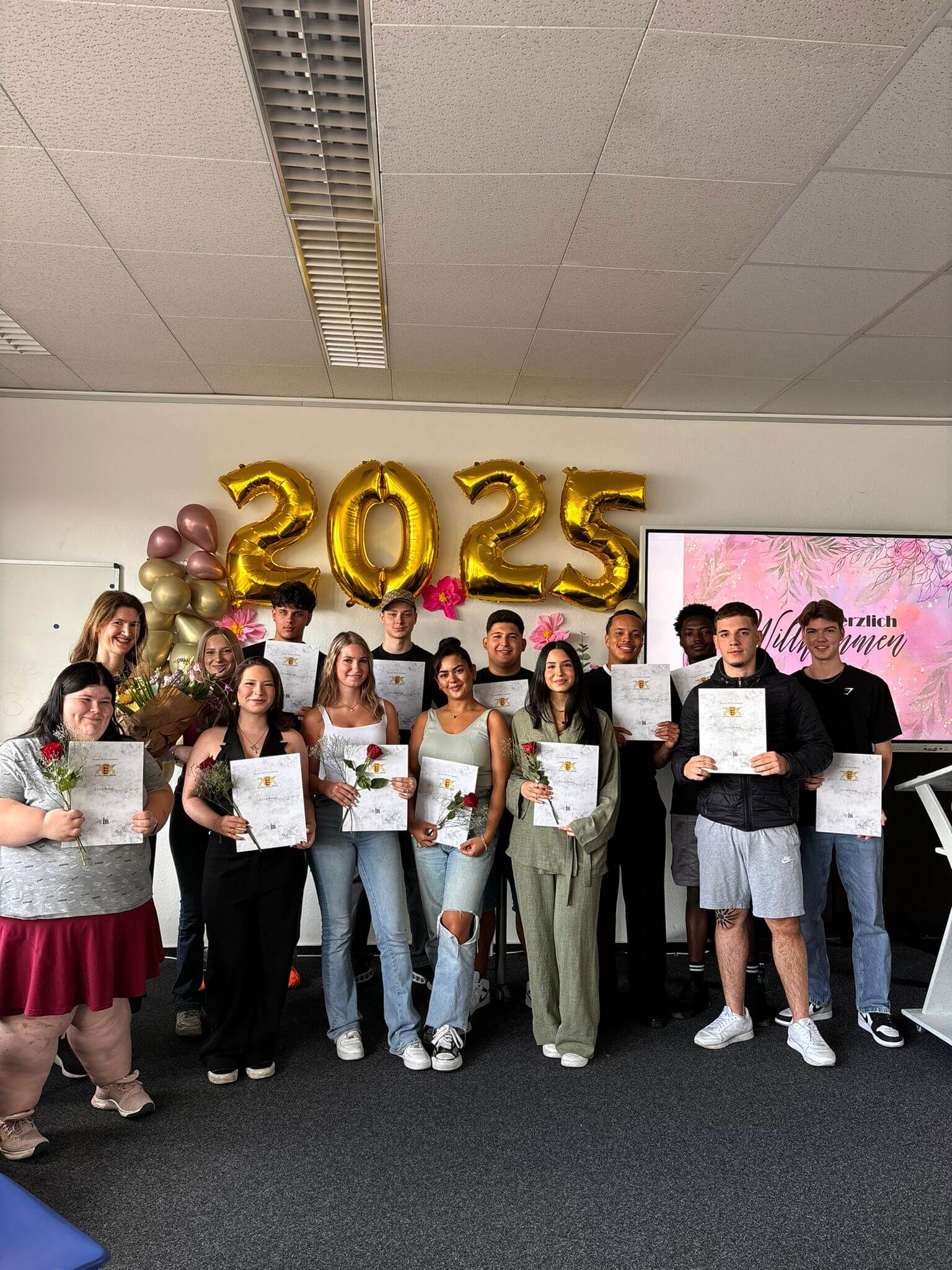 Eine Gruppe von jungen Menschen hält Zertifikate in der Hand, während goldene Luftballons die Zahl 2025 zeigen.